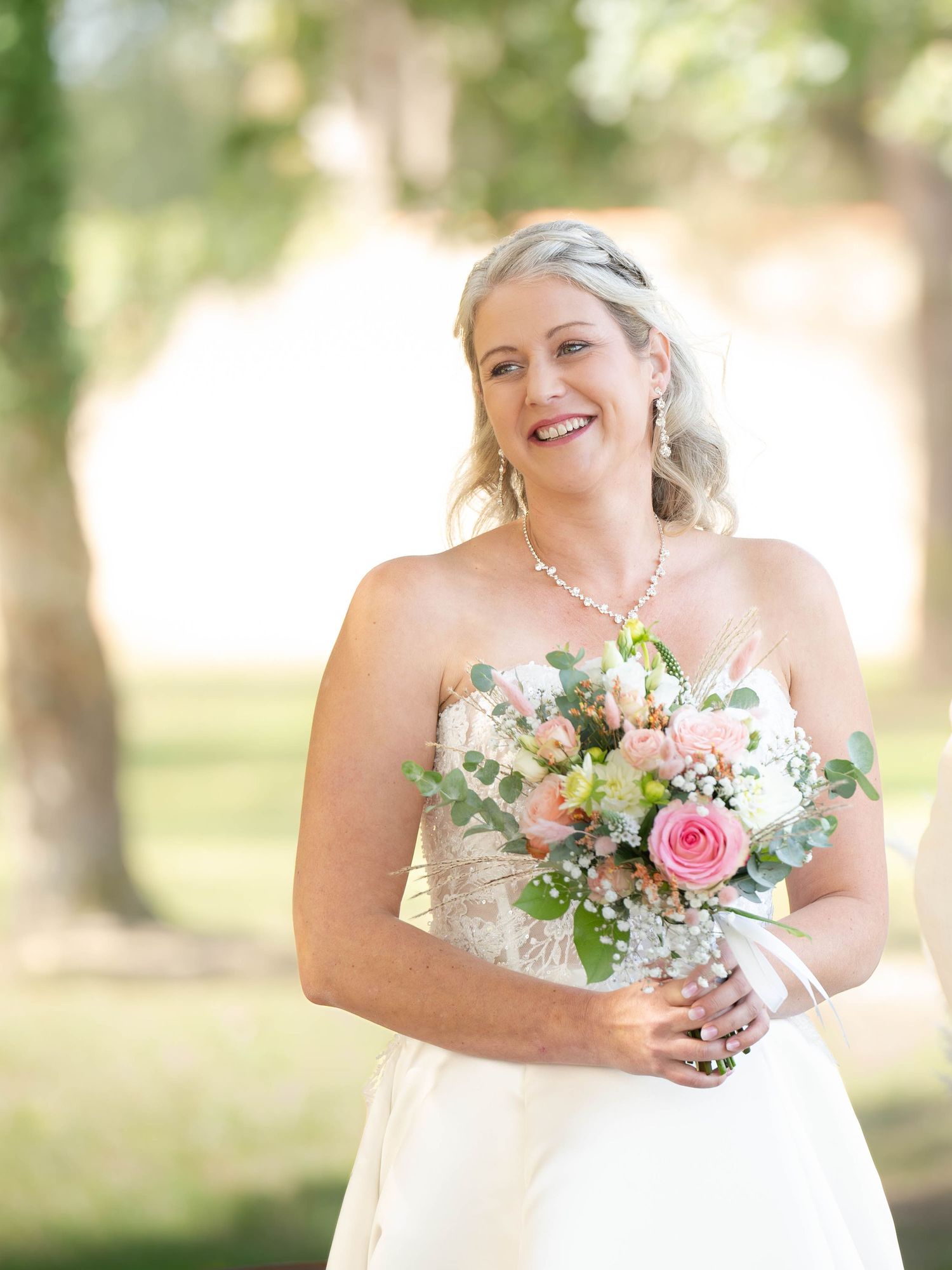 A bride in a white strapless gown holds a pink and white floral bouquet while posing outdoors in a sunlit garden setting.