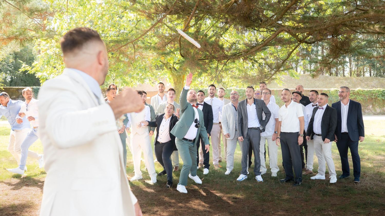 A group of well-dressed guests gather outdoors under a large tree during a daytime celebration event.