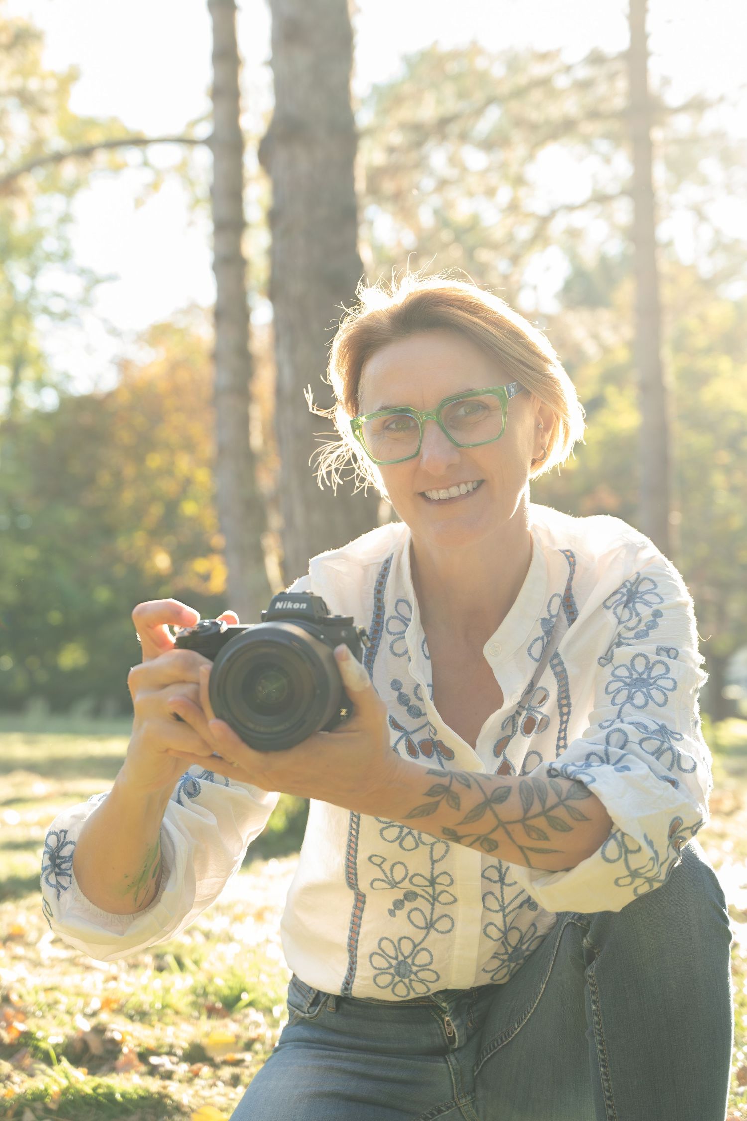 Photographer in white and blue patterned shirt takes photos with professional camera in golden sunlit forest.