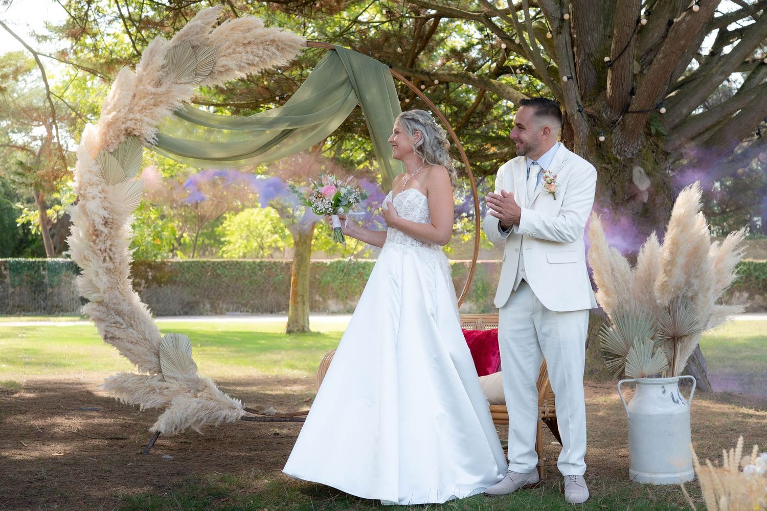 A couple in white outfits poses for romantic wedding photos under trees in a sunlit park setting.