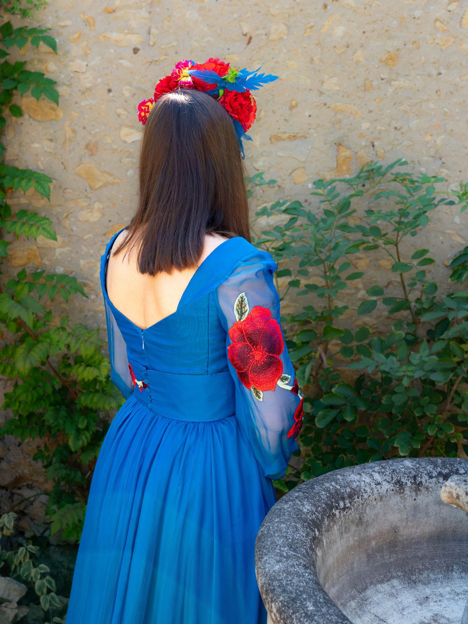 Back view of blue dress with sheer sleeves against stone wall and greenery.