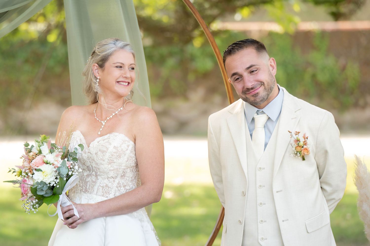 A bride and groom pose together outdoors in white wedding attire with a bouquet of flowers.