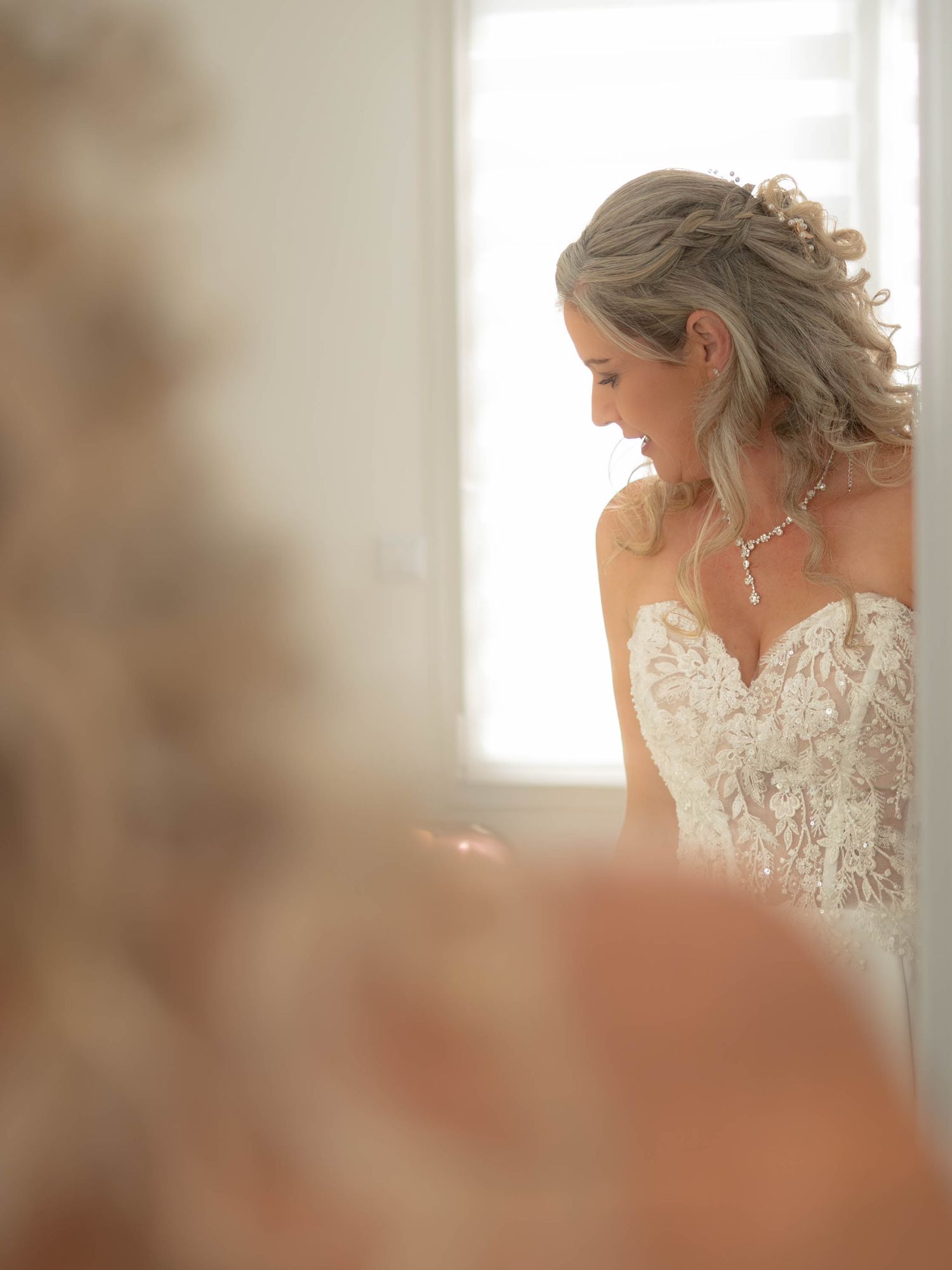 Bride in delicate lace strapless wedding gown stands in soft window light wearing elegant necklace and upswept hairstyle.