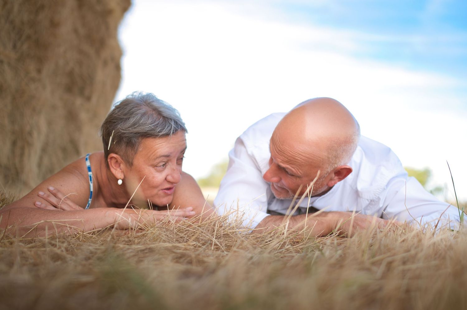 Two people resting together on dried grass against a blue summer sky.