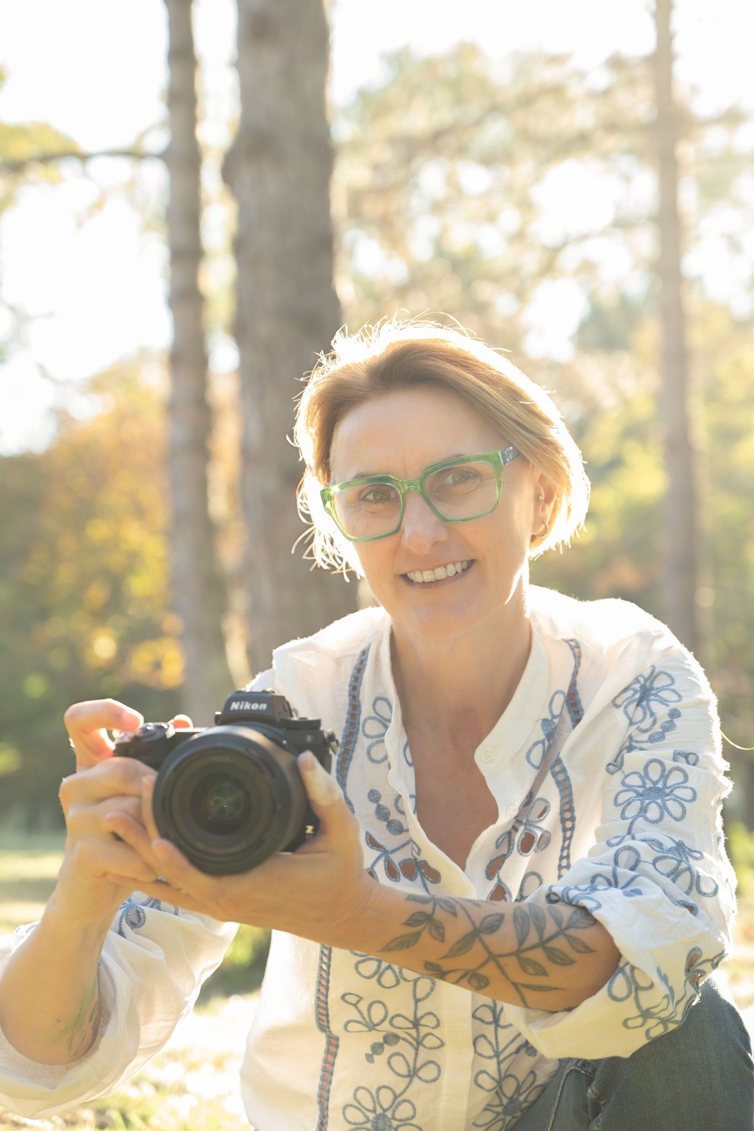 A photographer in a white embroidered top holds up a camera while smiling in sunlit outdoor setting.