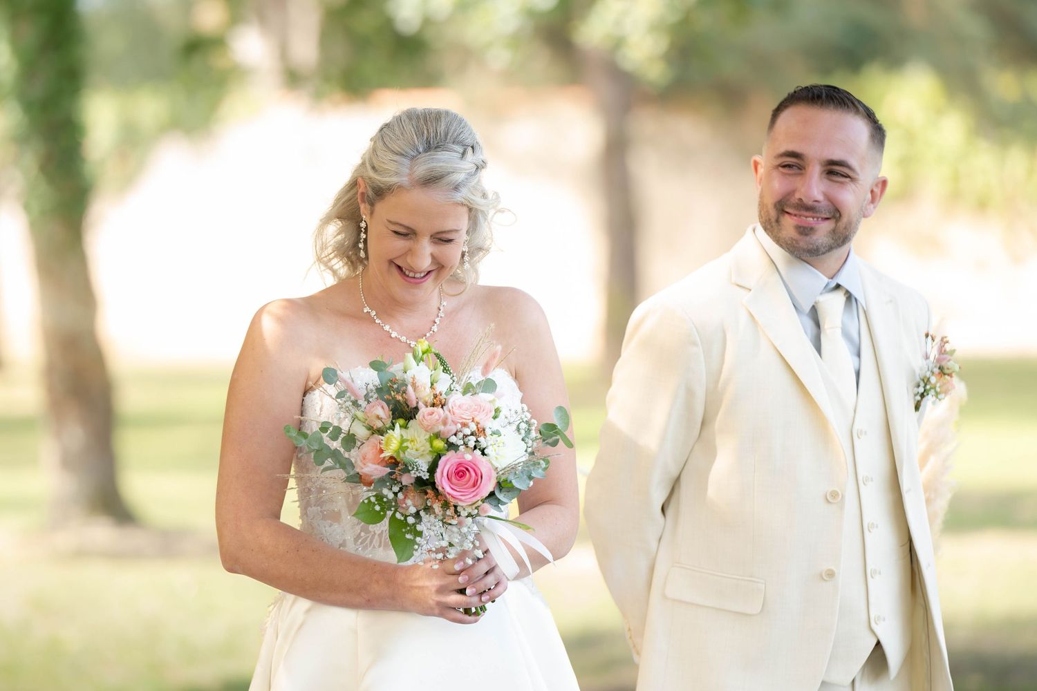 A wedding couple stands outdoors in natural lighting with the bride holding a pink and white floral bouquet.