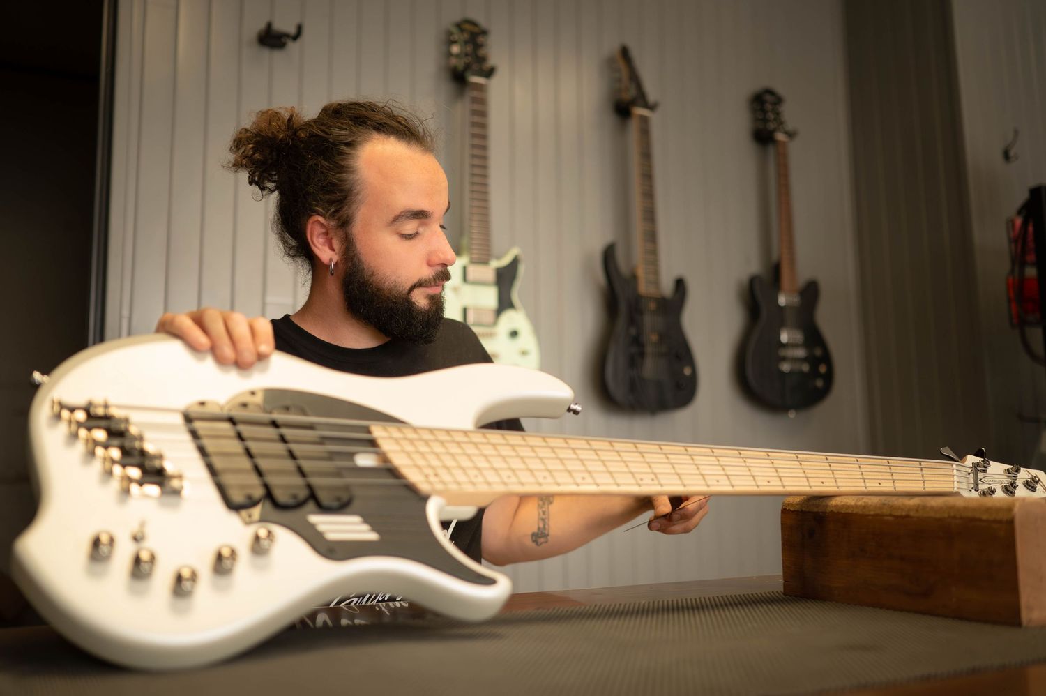 Electric bass guitar being examined in workshop with guitars displayed on wall.