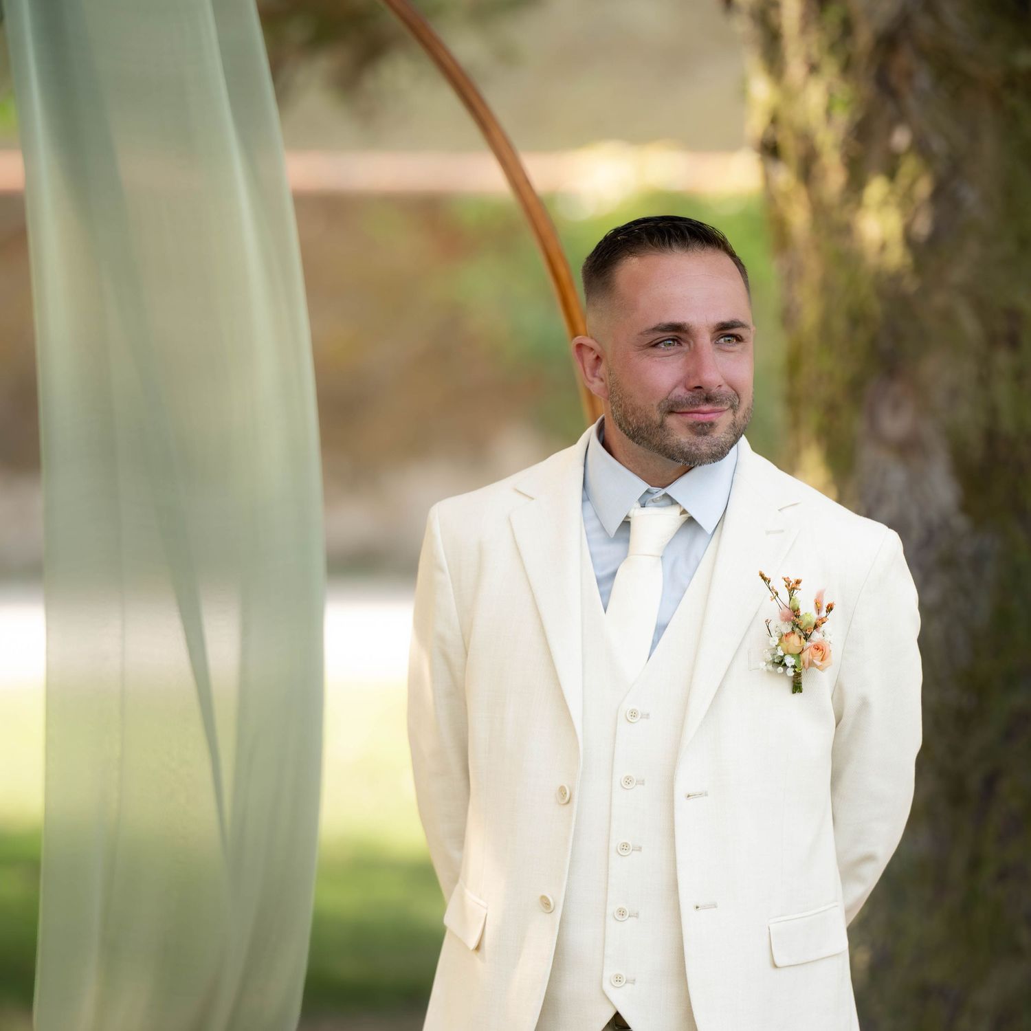 A groom in a crisp white tuxedo stands outdoors during a wedding ceremony.