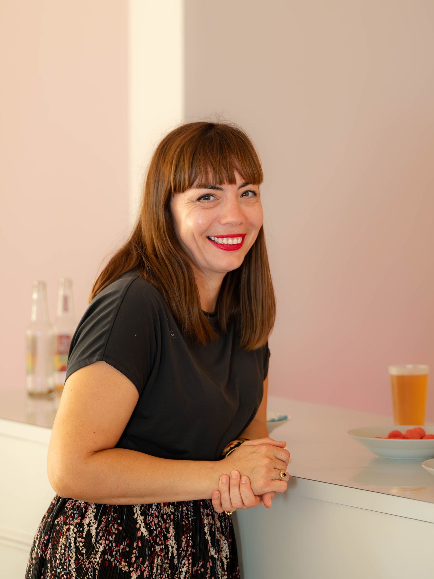 A person in a black top and patterned skirt leans against a pink counter in a bright modern space.