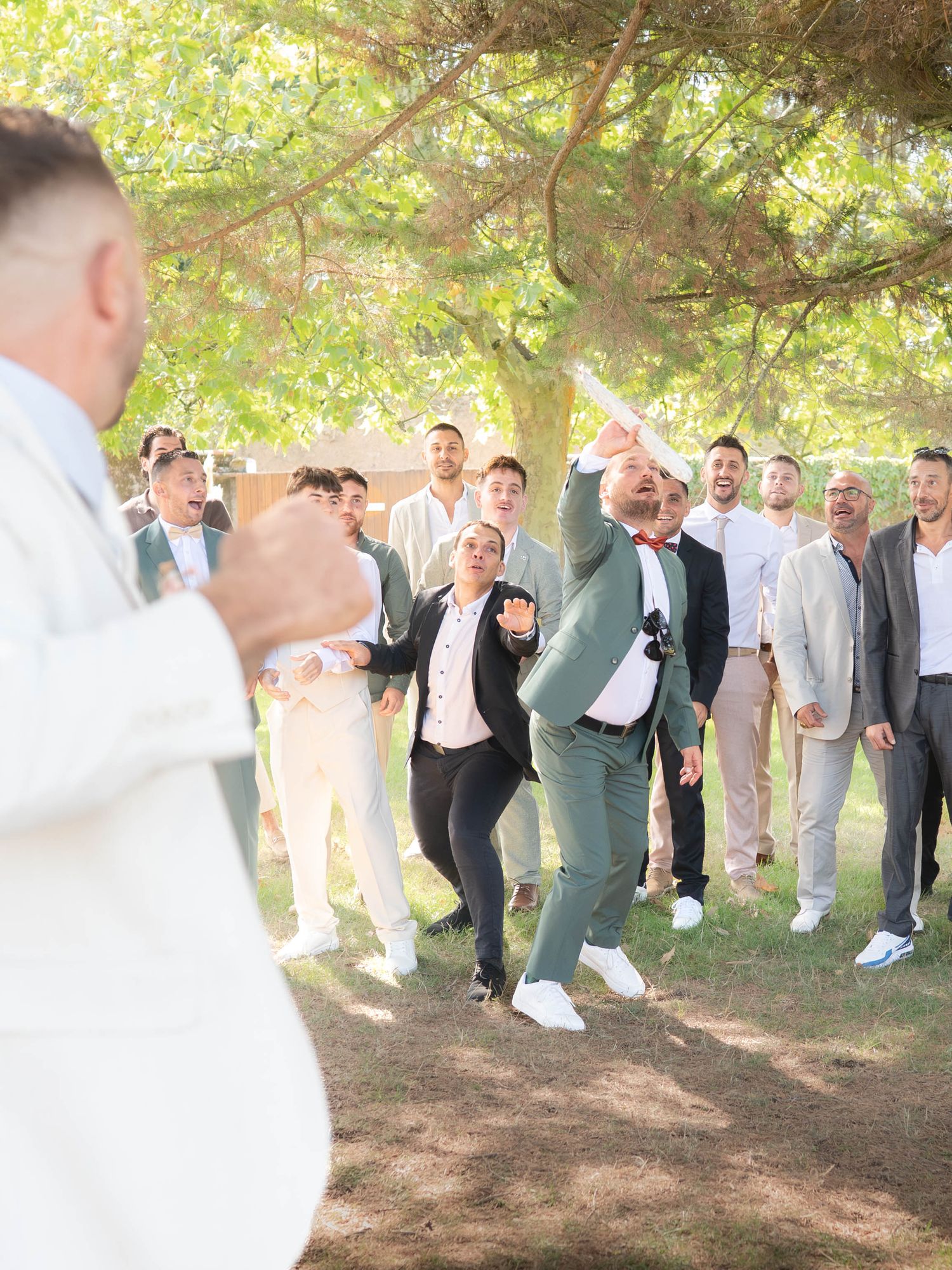 Wedding party members lined up under tree branches performing a synchronized dance move at an outdoor celebration.