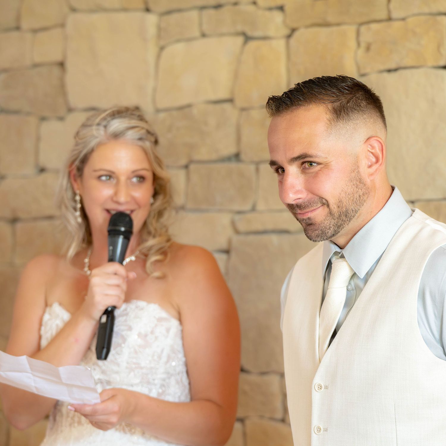 A couple at their wedding ceremony with the bride speaking into a microphone against a stone wall backdrop.