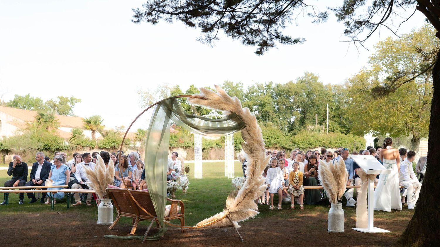 Outdoor wedding ceremony under a circular arch with guests seated on white chairs in a garden setting.