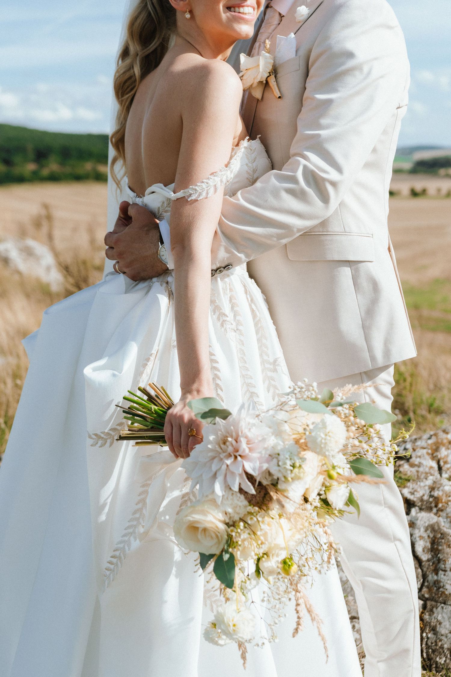 Romantic wedding couple embracing in a sunlit field with a delicate white bridal bouquet.