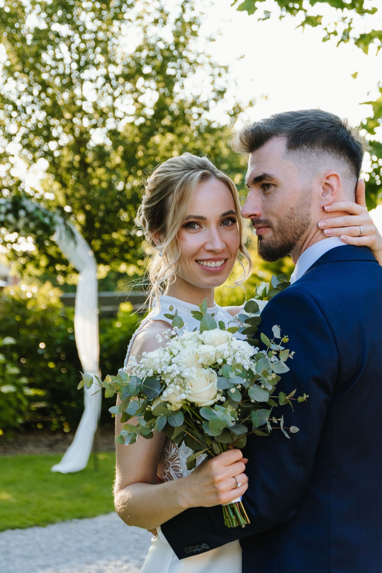 Newlywed couple with white floral bouquet posing outdoors.