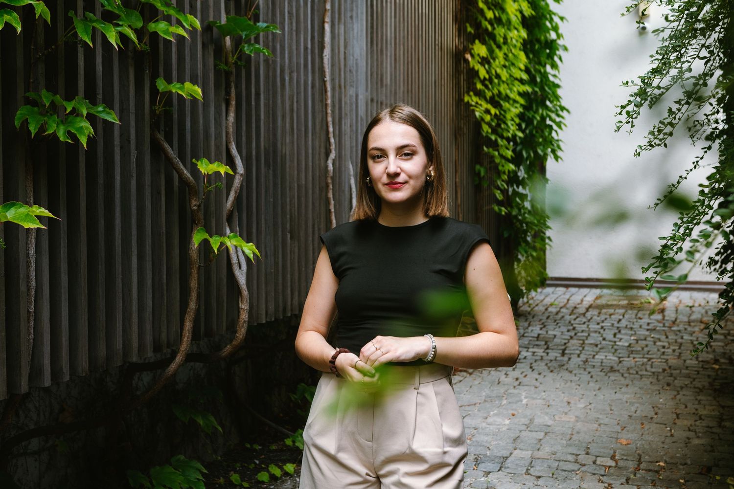 A woman wearing a black top and beige pants stands casually by a wooden wall with green plants.