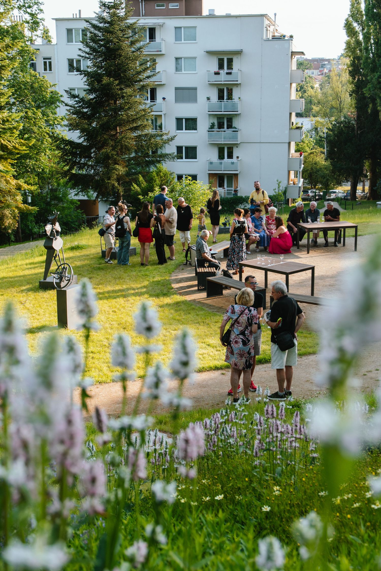 Group of people gathered outside apartment buildings with wildflowers in the foreground.