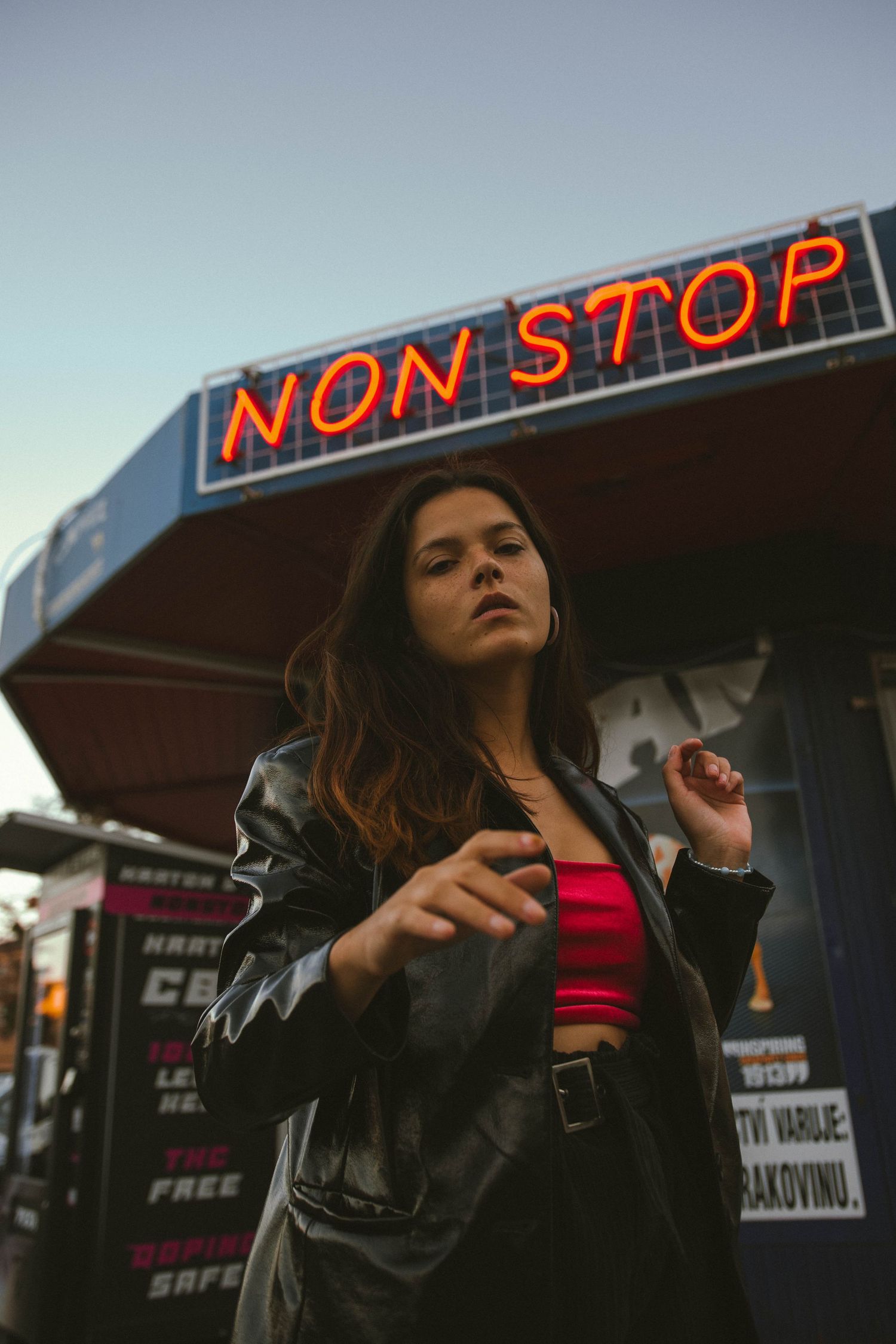 A person in leather jacket and red top poses beneath a neon Non Stop sign at dusk.