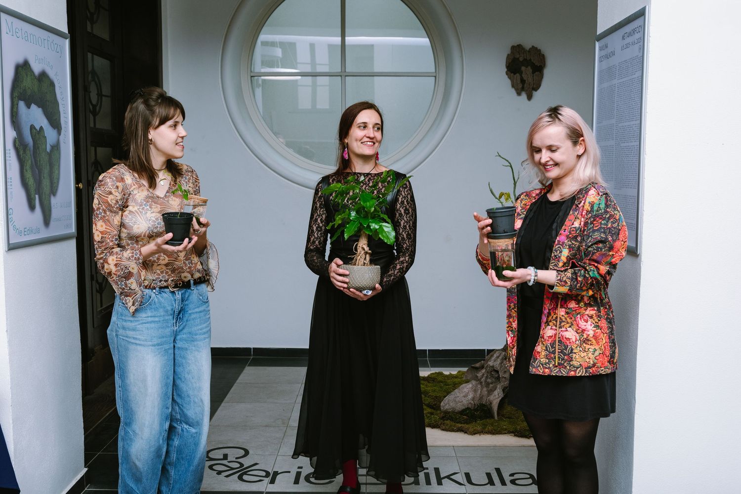 Three people holding potted plants while standing in front of a circular window.