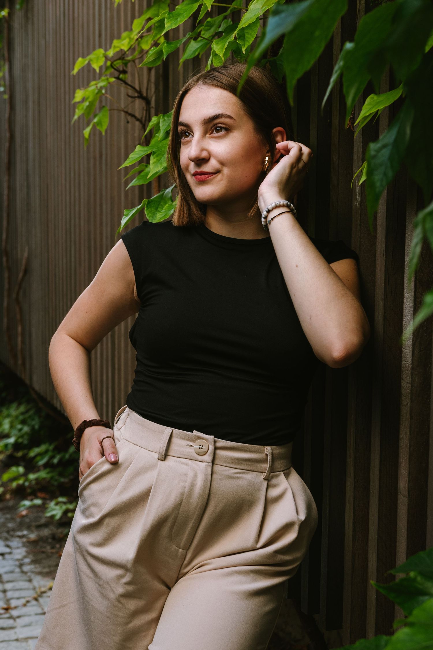 A woman in a black sleeveless top and beige pants poses against a wooden fence surrounded by green foliage.
