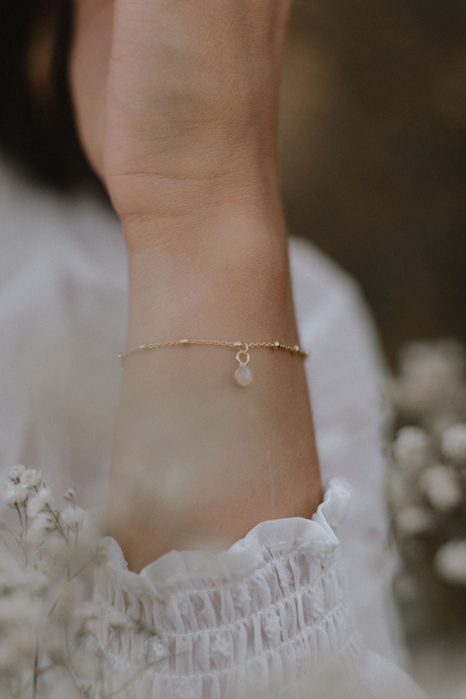 Delicate gold chain bracelet with small charm worn on wrist with ruffled white sleeve detail.