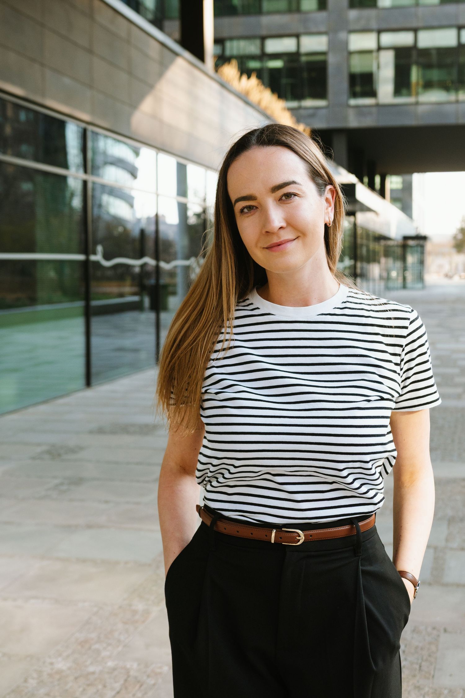 Person wearing striped shirt and dark pants poses against modern architecture.
