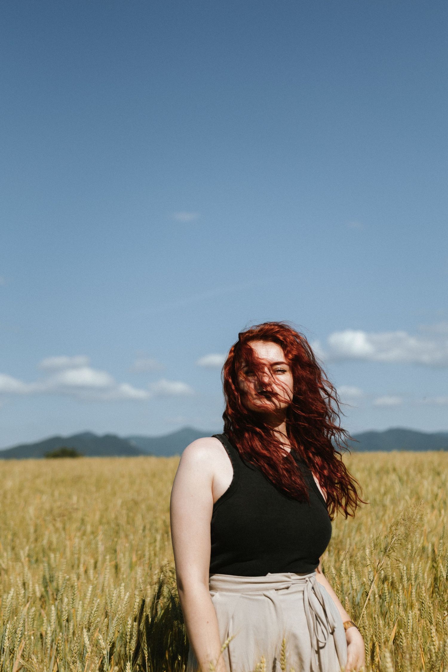 A woman with flowing red hair stands in a golden wheat field on a sunny day wearing a dark top and light skirt.