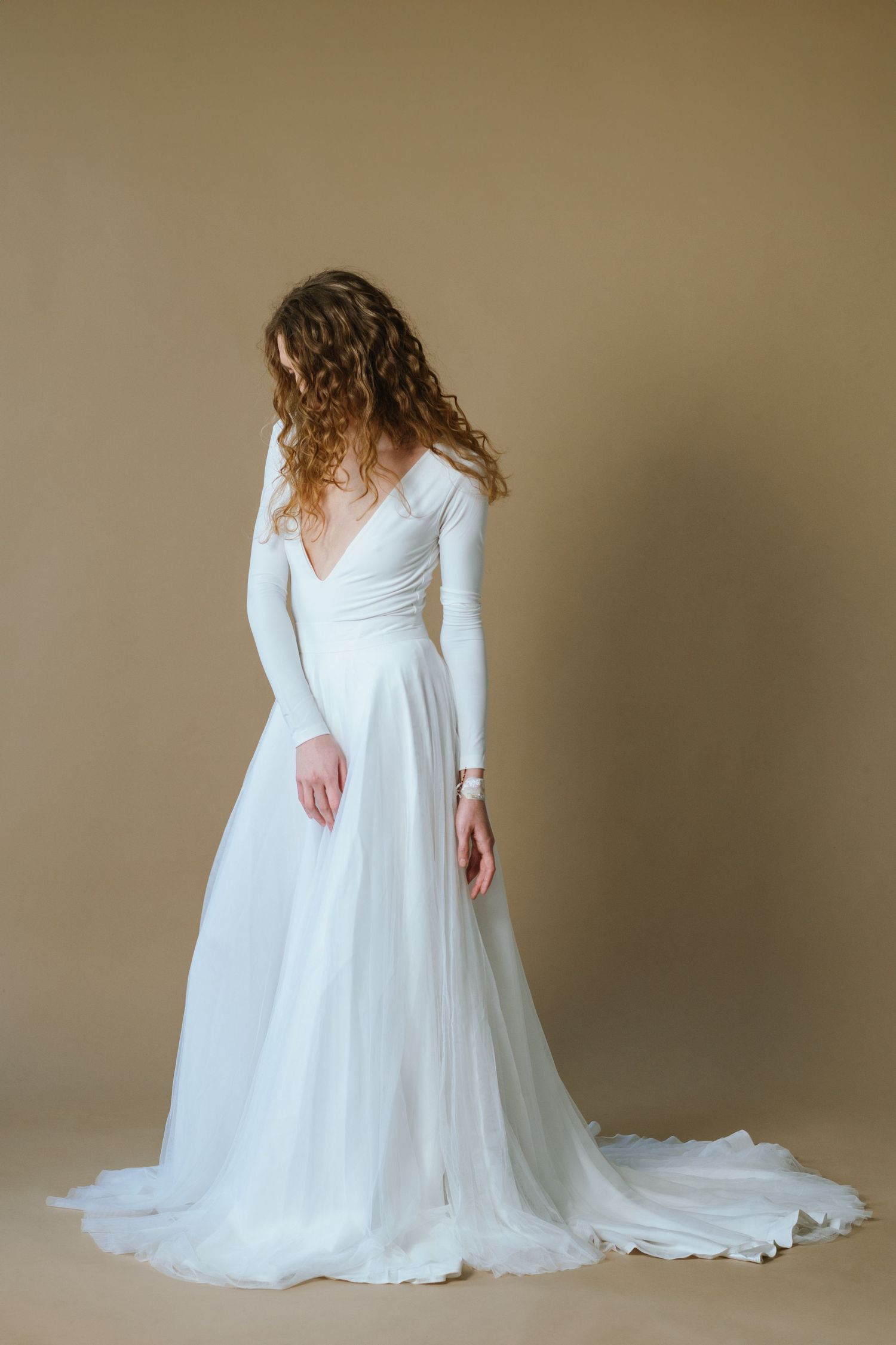 Elegant long-sleeved white wedding gown with flowing train photographed against beige backdrop.