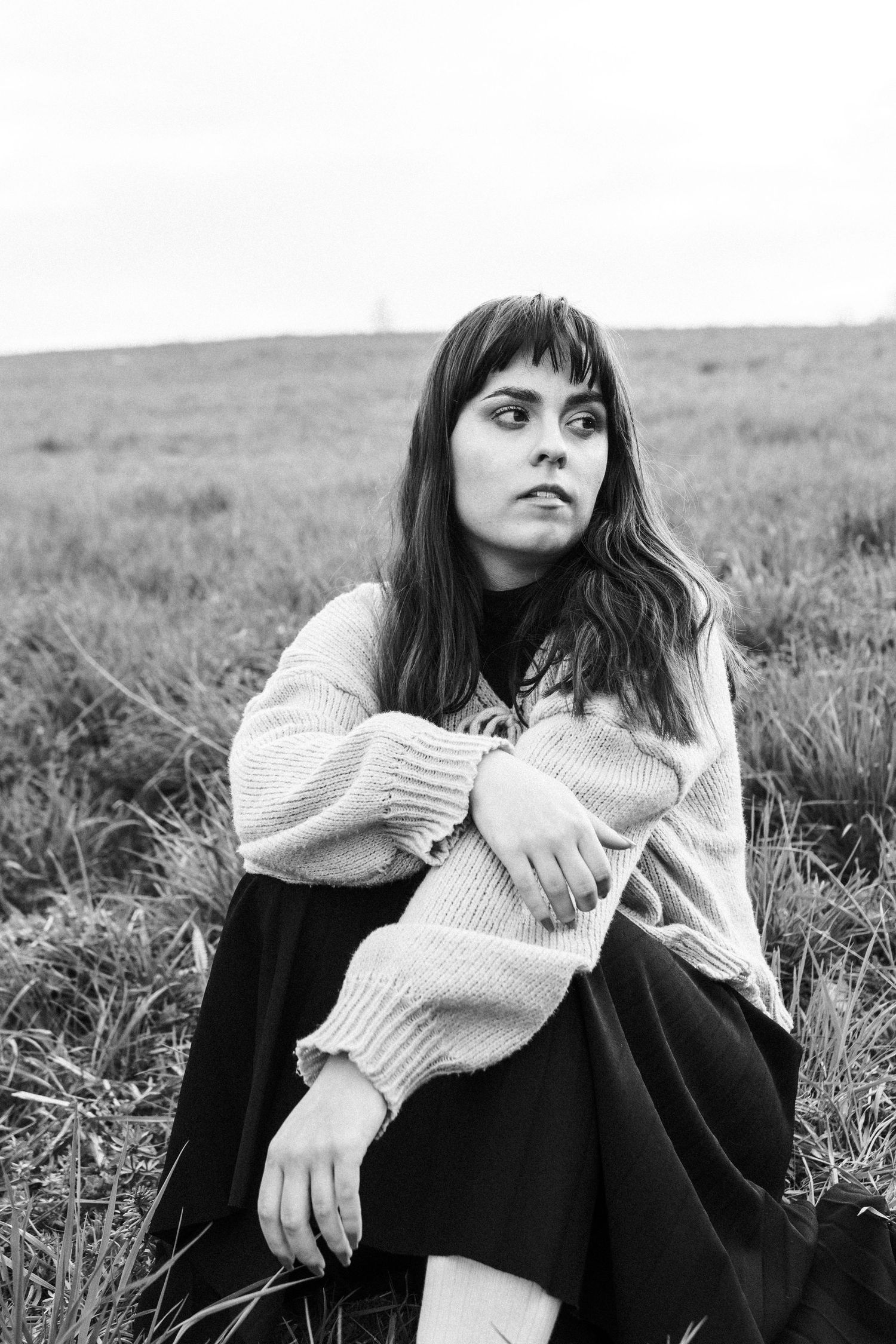 Person in sweater and skirt sits thoughtfully in black and white countryside portrait.