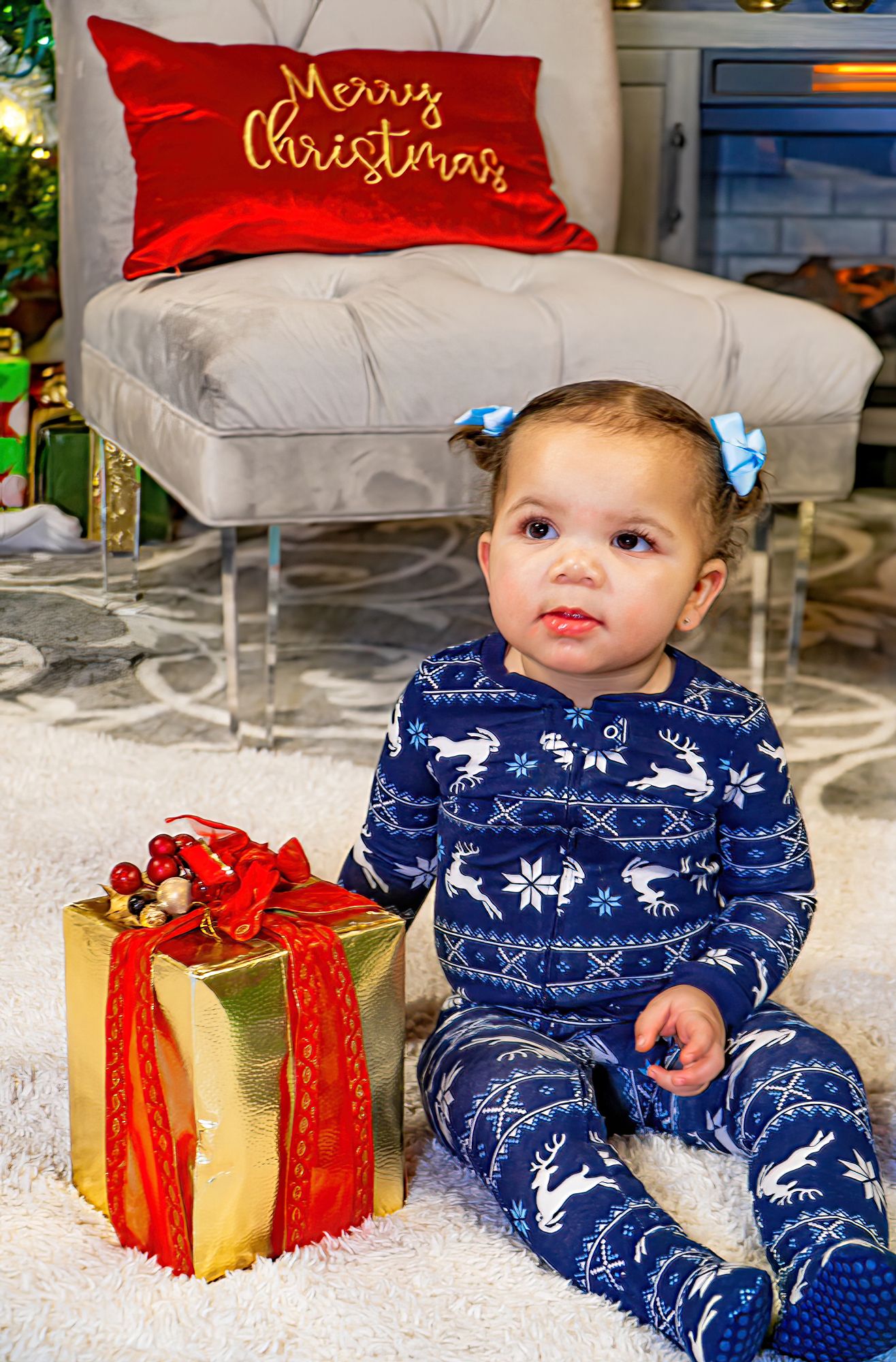 Baby girl in blue reindeer pajamas sitting beside gold wrapped Christmas gift.