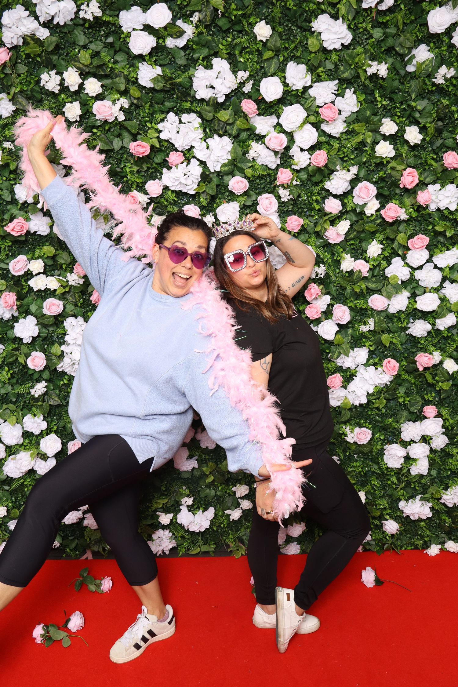 Two women pose playfully with a pink feather boa and heart sunglasses in front of a floral wall backdrop on a red carpet.
