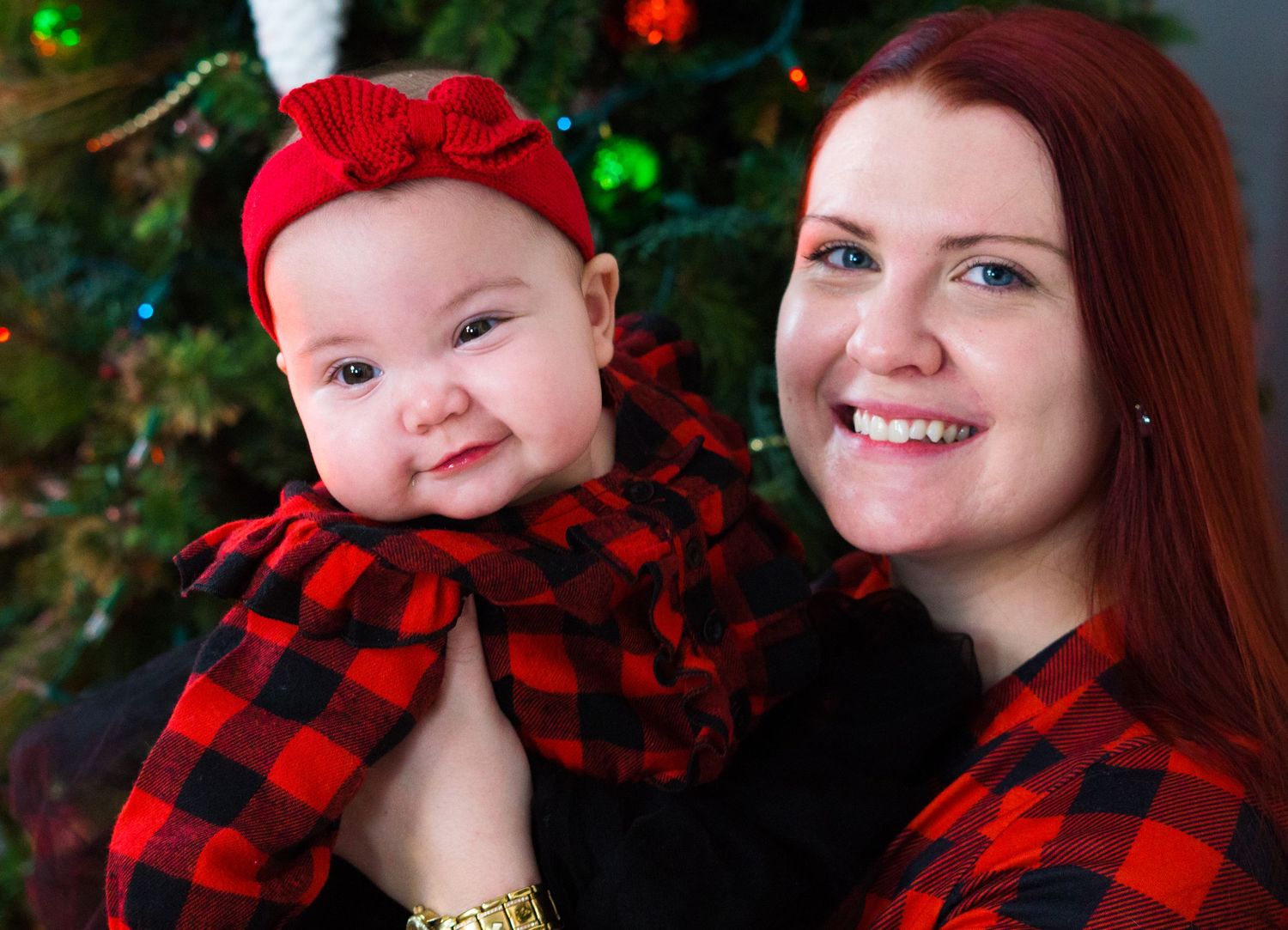 Smiling mother with red hair holds adorable baby in matching red plaid outfits before Christmas tree.