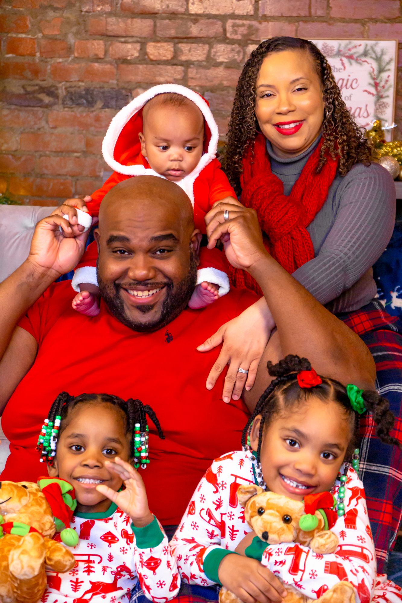 Smiling family of five in festive red Christmas outfits poses together with baby in Santa costume.