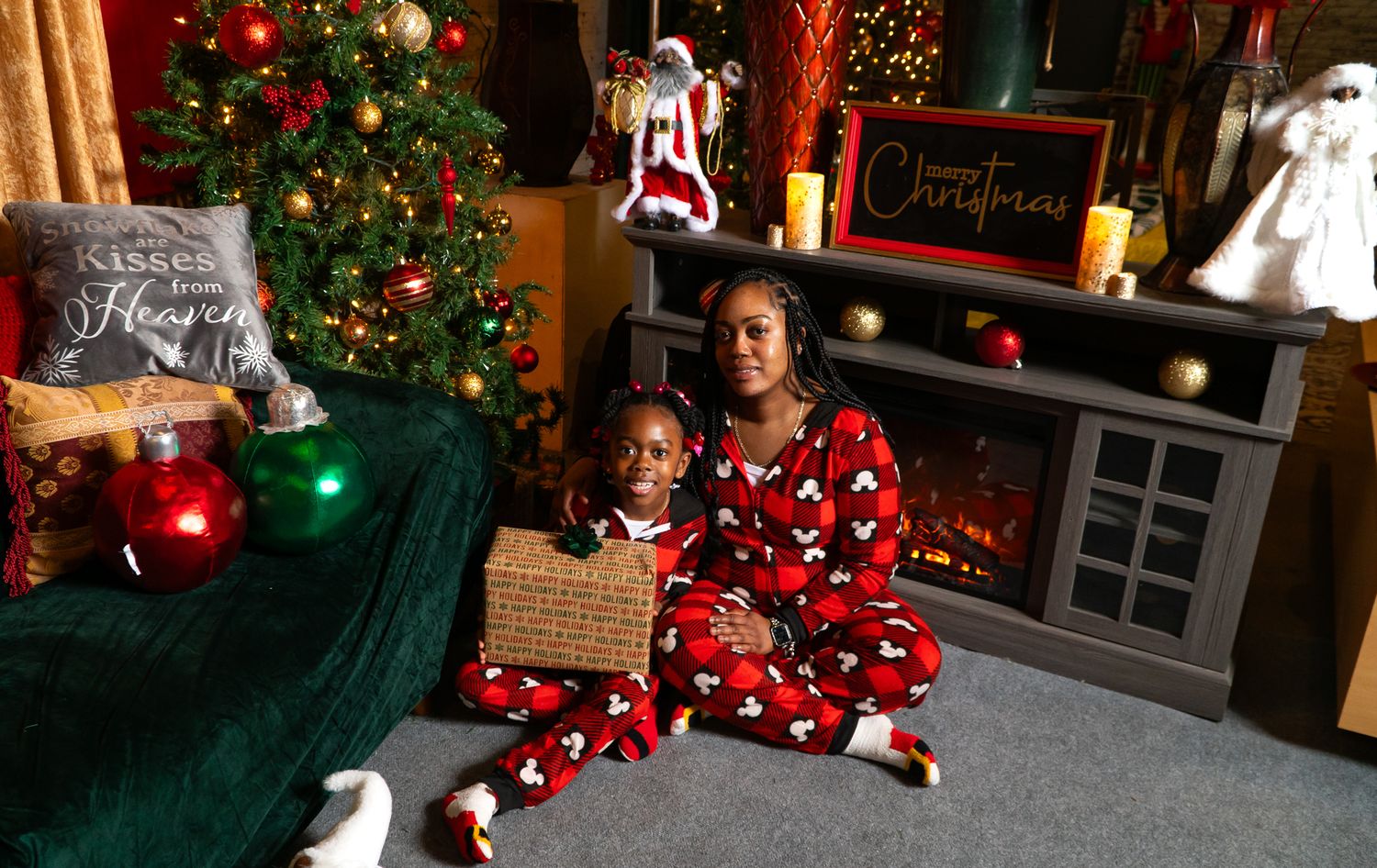 Mother and child in matching red Mickey Mouse pajamas sitting together near a decorated Christmas tree and fireplace.