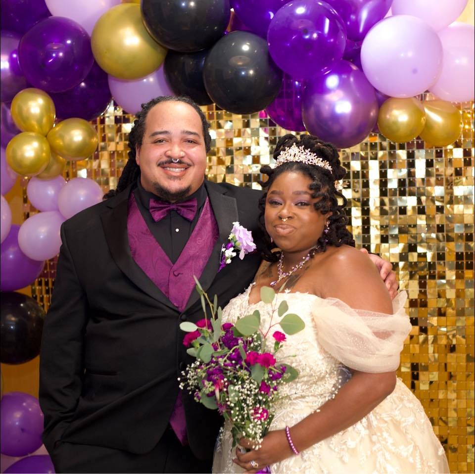 Couple posing at wedding with purple, gold, and black balloon arch backdrop.