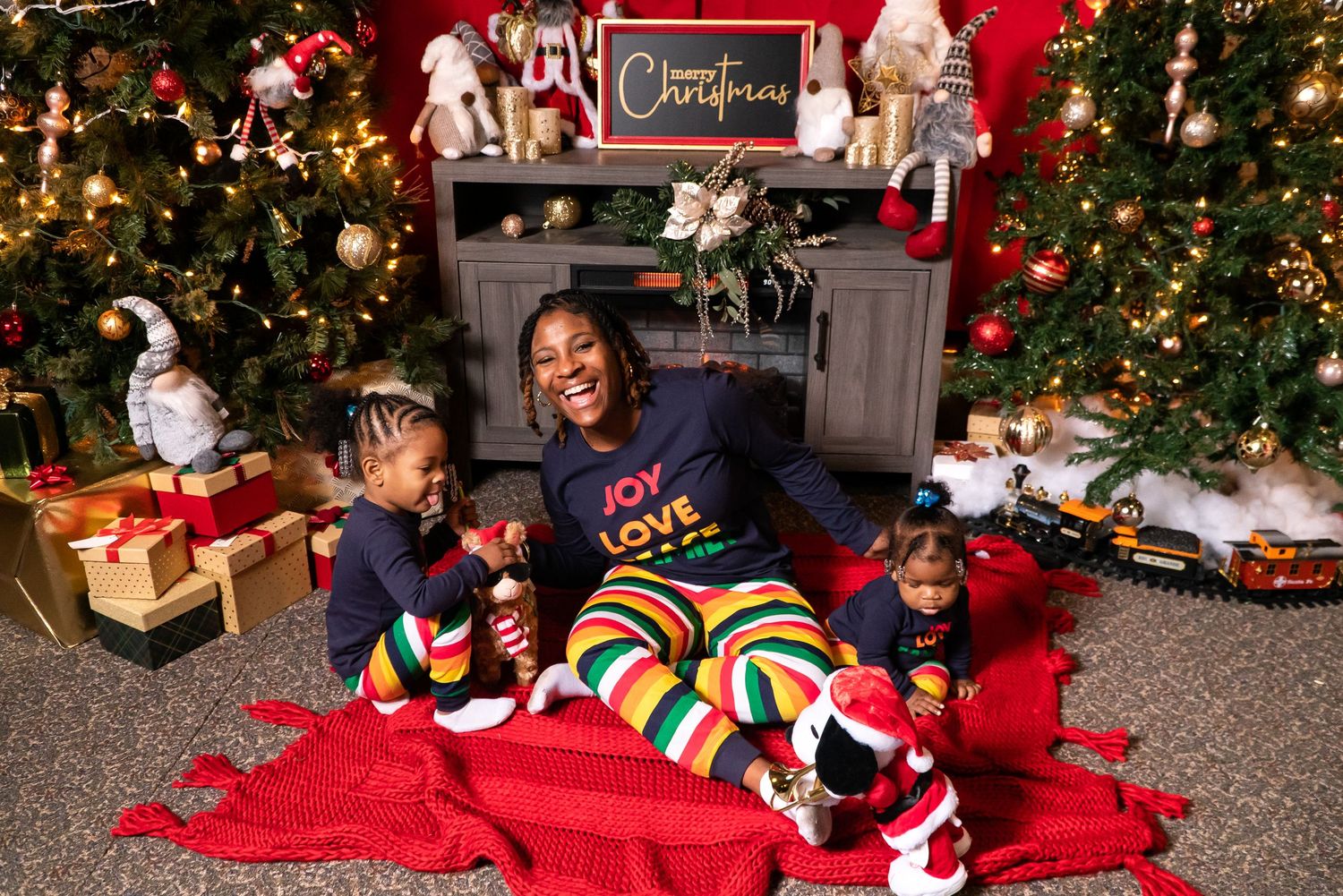 Joyful mother in holiday pajamas sitting with two young children by Christmas fireplace.