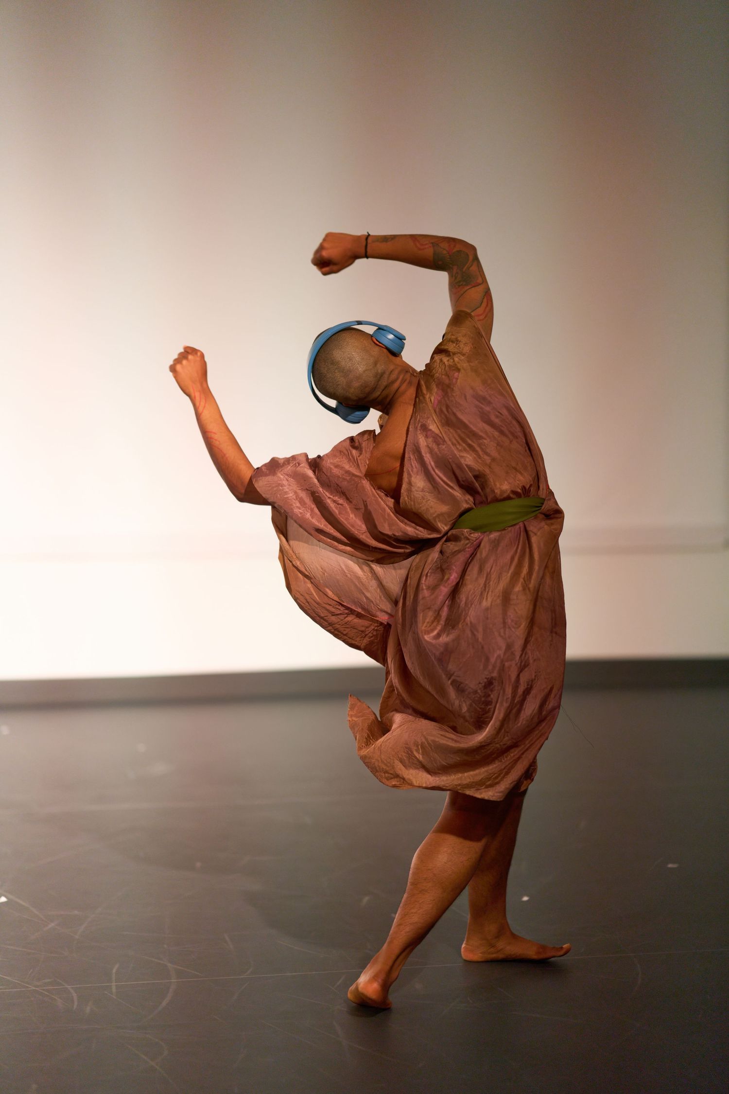 A barefoot dancer in a flowing brown costume and blue headphones performs an expressive modern dance pose on stage.