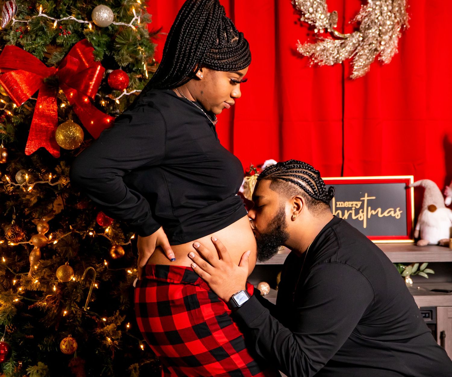 Couple shares an intimate Christmas moment as man kisses pregnant partners baby bump by a decorated tree.