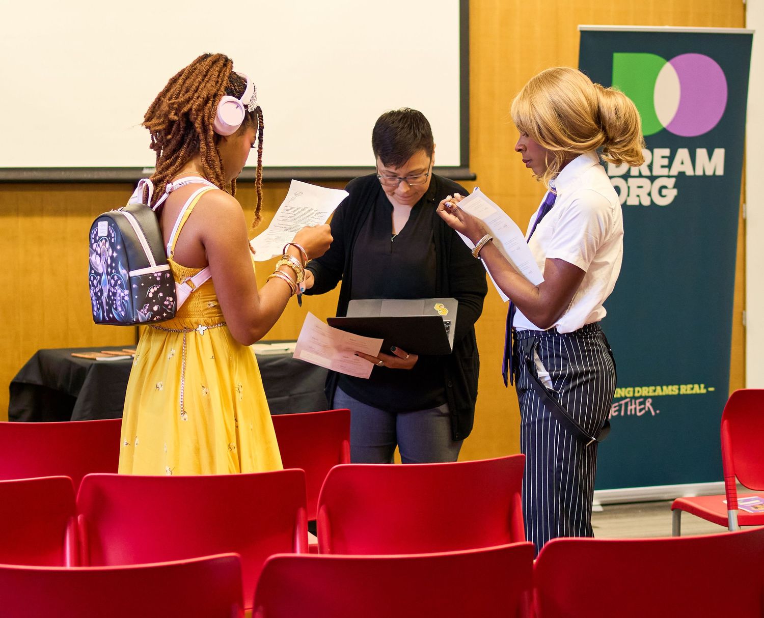 Three women reviewing documents at a Dream.org event, standing among red chairs in a conference room.
