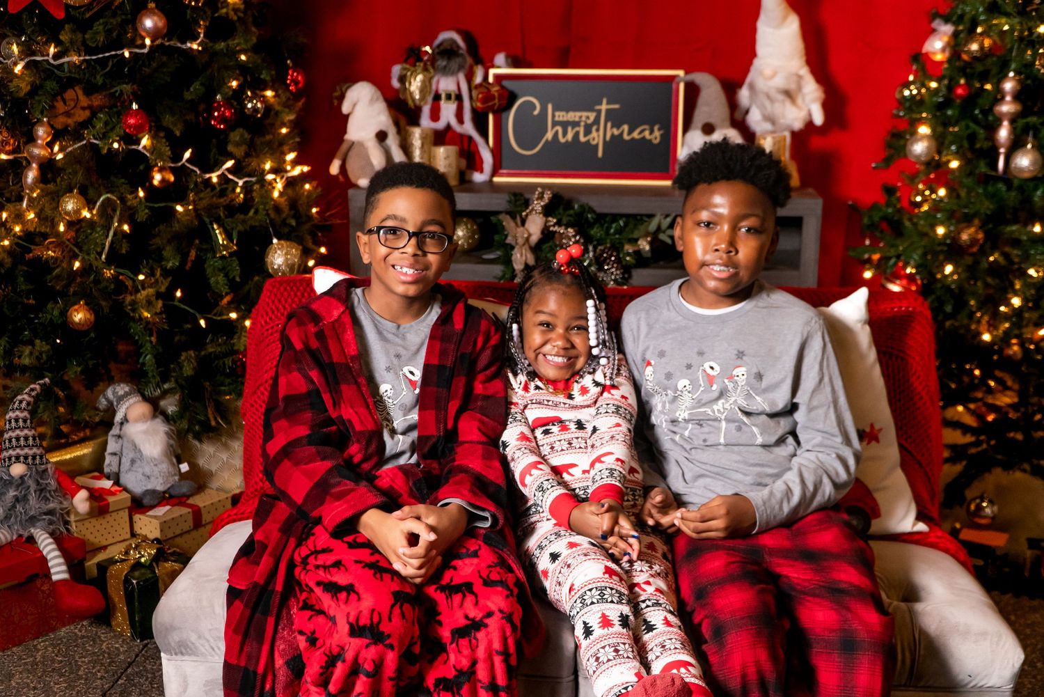 Three children pose together in Christmas pajamas beside a decorated holiday tree.