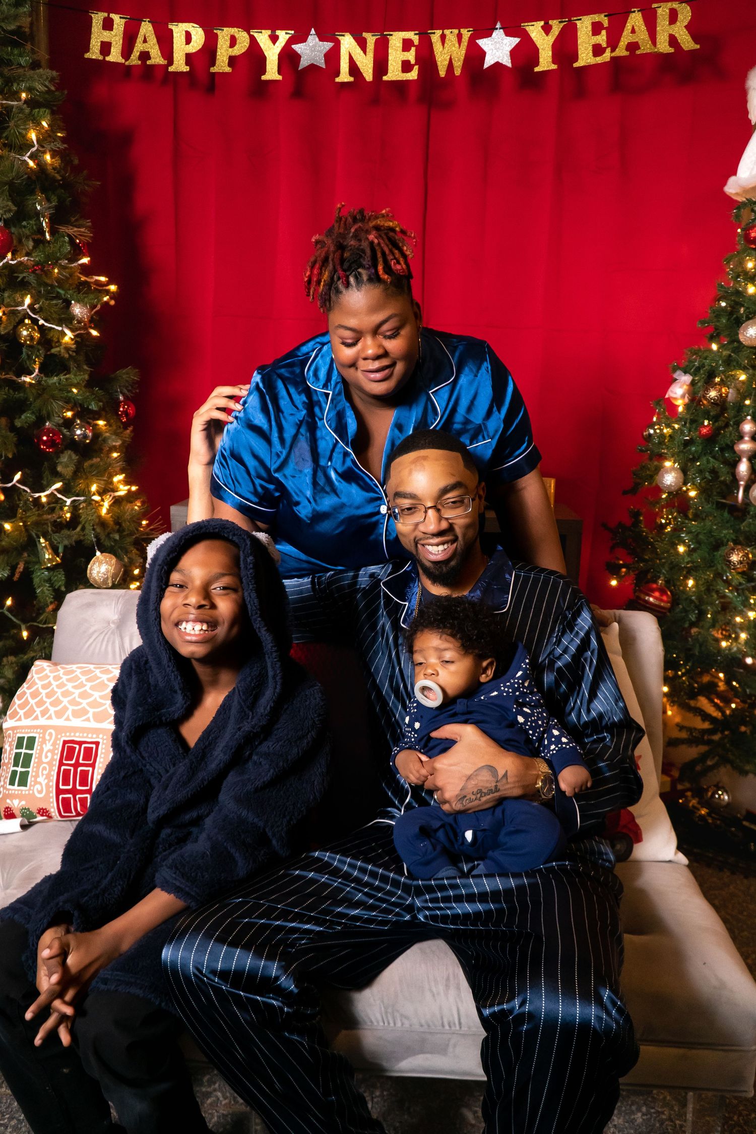 Family of four in blue satin pajamas posing under a Happy New Year banner with Christmas trees.
