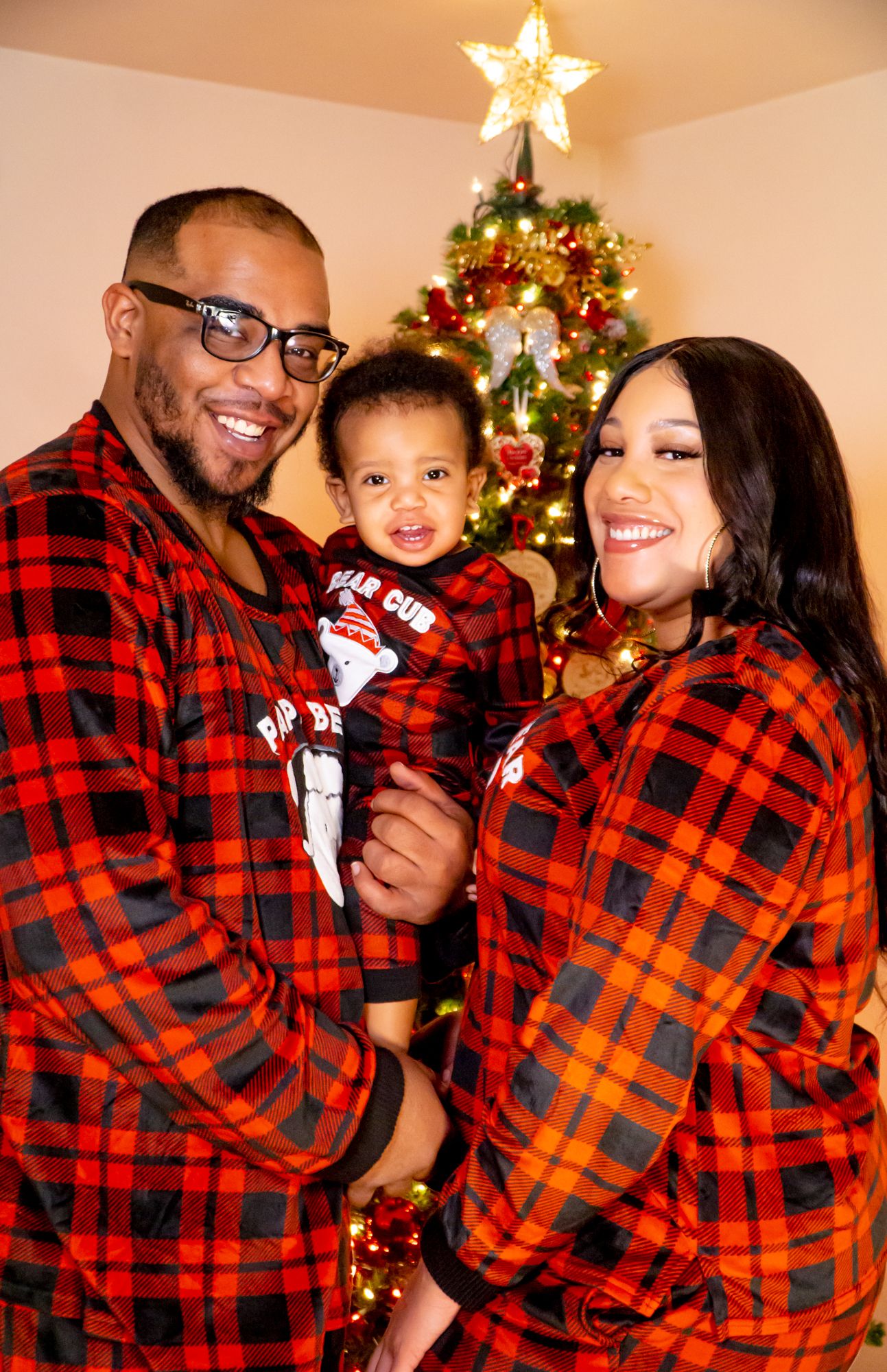 Family of three in matching red plaid pajamas posing together by decorated Christmas tree.