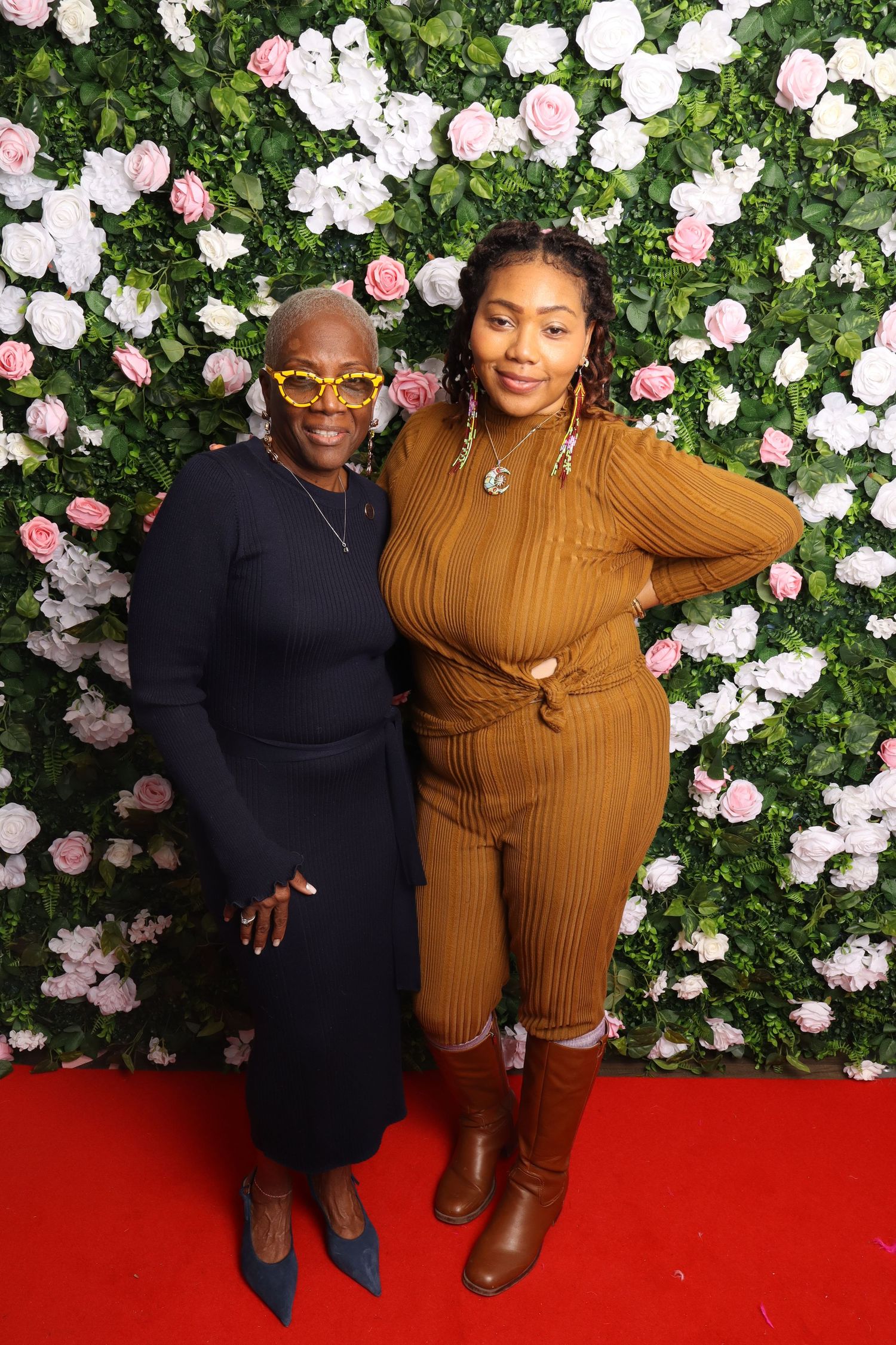 Two women pose together in front of a floral wall backdrop on a red carpet at a formal event.