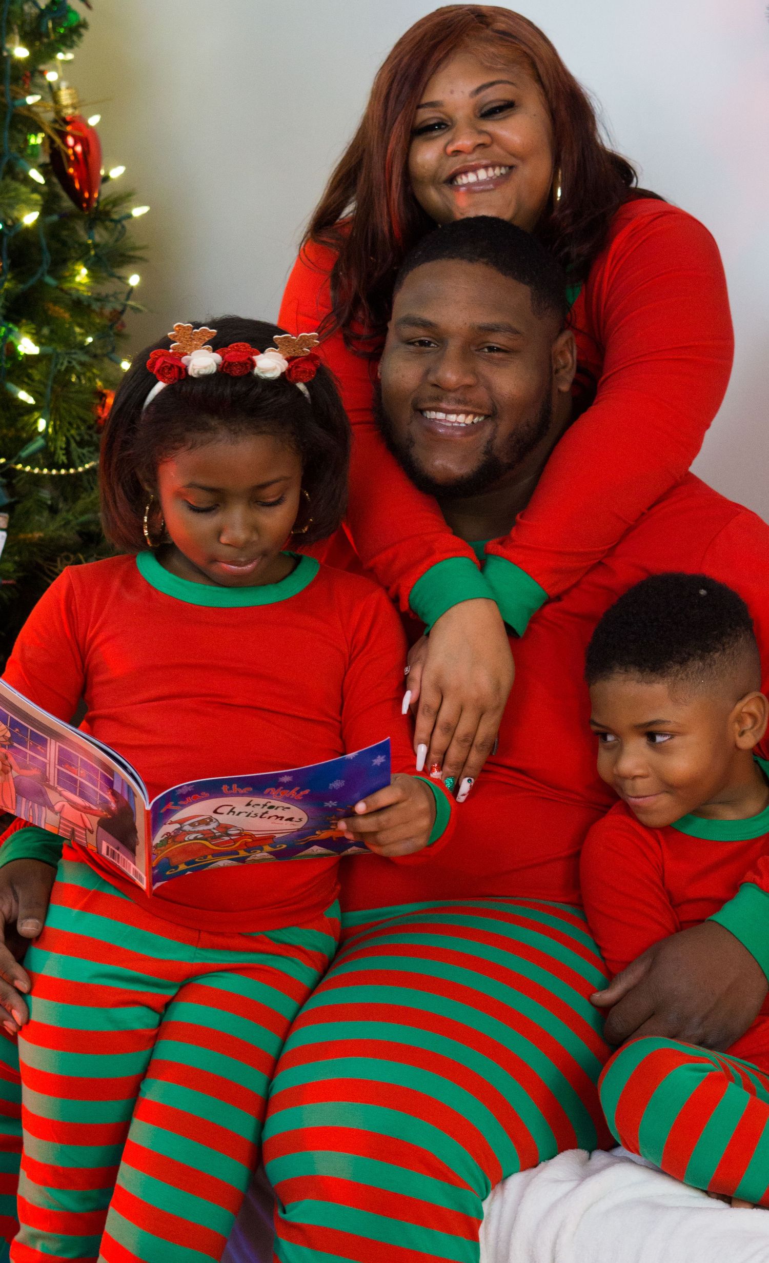 Family in red and green striped Christmas pajamas sits together by tree, girl reading a Christmas book.