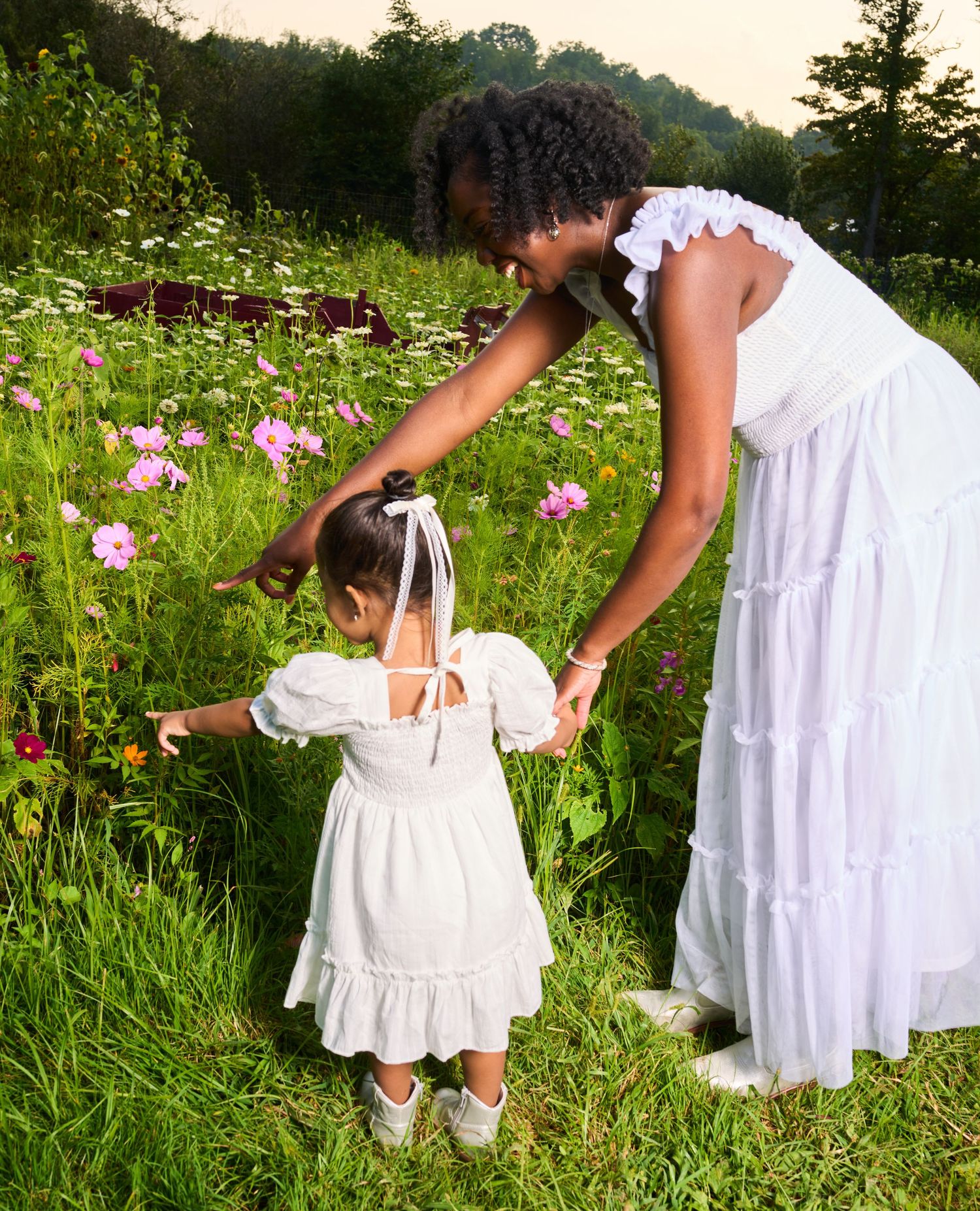 A mother in a white dress holds her toddler daughter's hand in a blooming wildflower meadow.