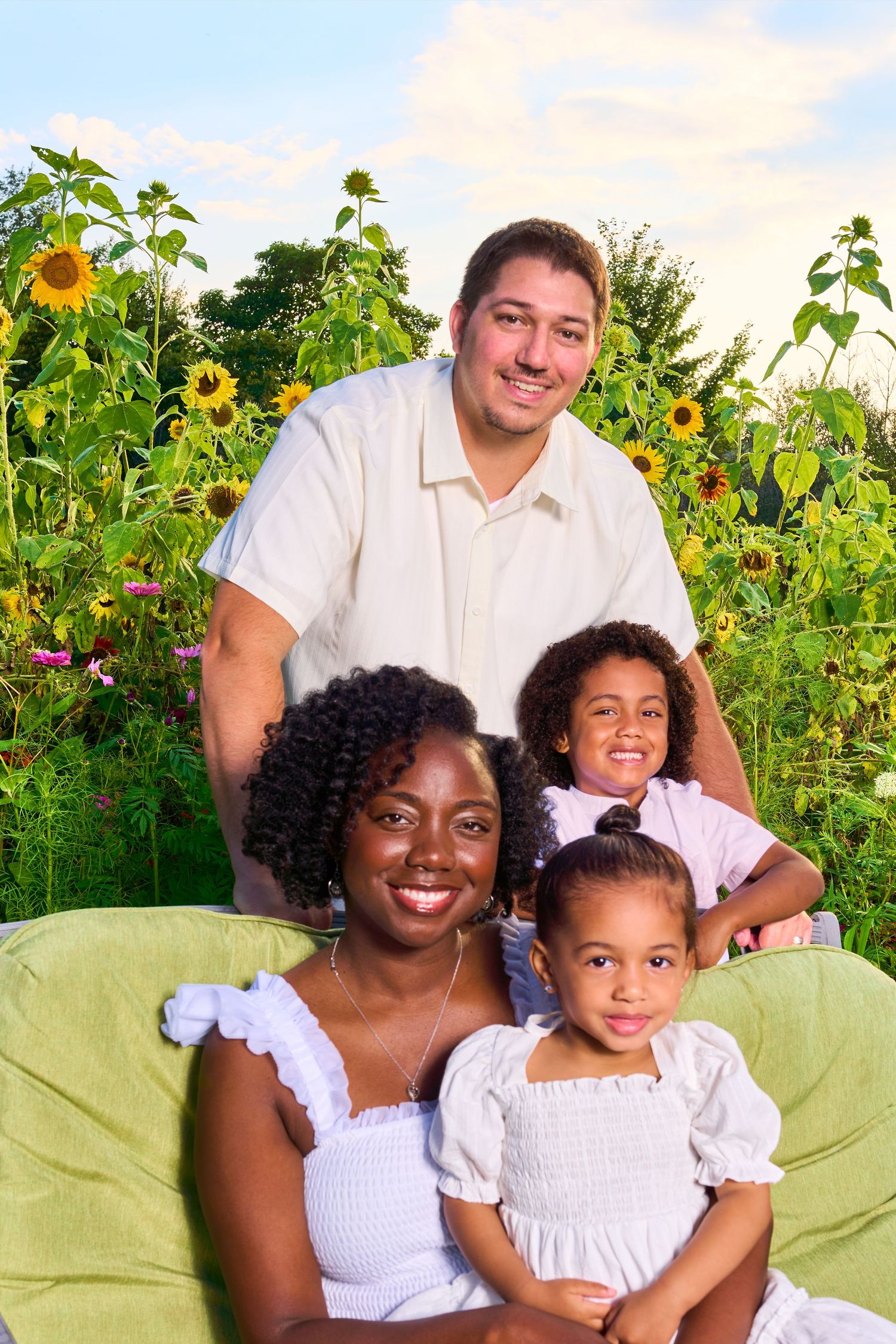 Interracial family of four smiling outdoors in a sunflower field, with two young daughters seated on a green chair.
