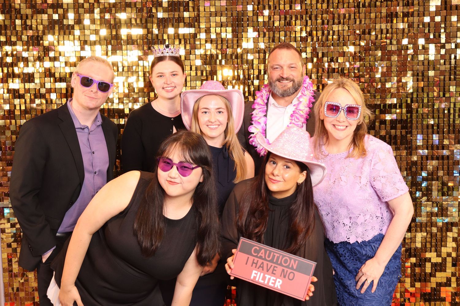 Group of seven friends posing at a glittery gold photo booth, wearing fun accessories including tiaras and sunglasses.