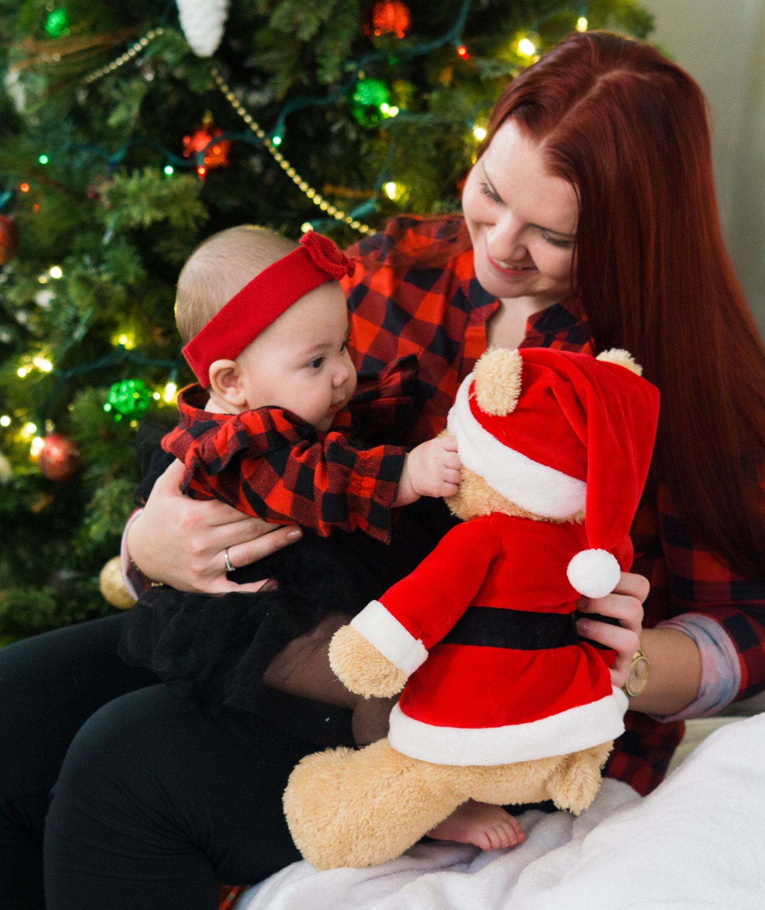 Mother holds baby and Santa teddy bear while sitting in front of a lit Christmas tree.
