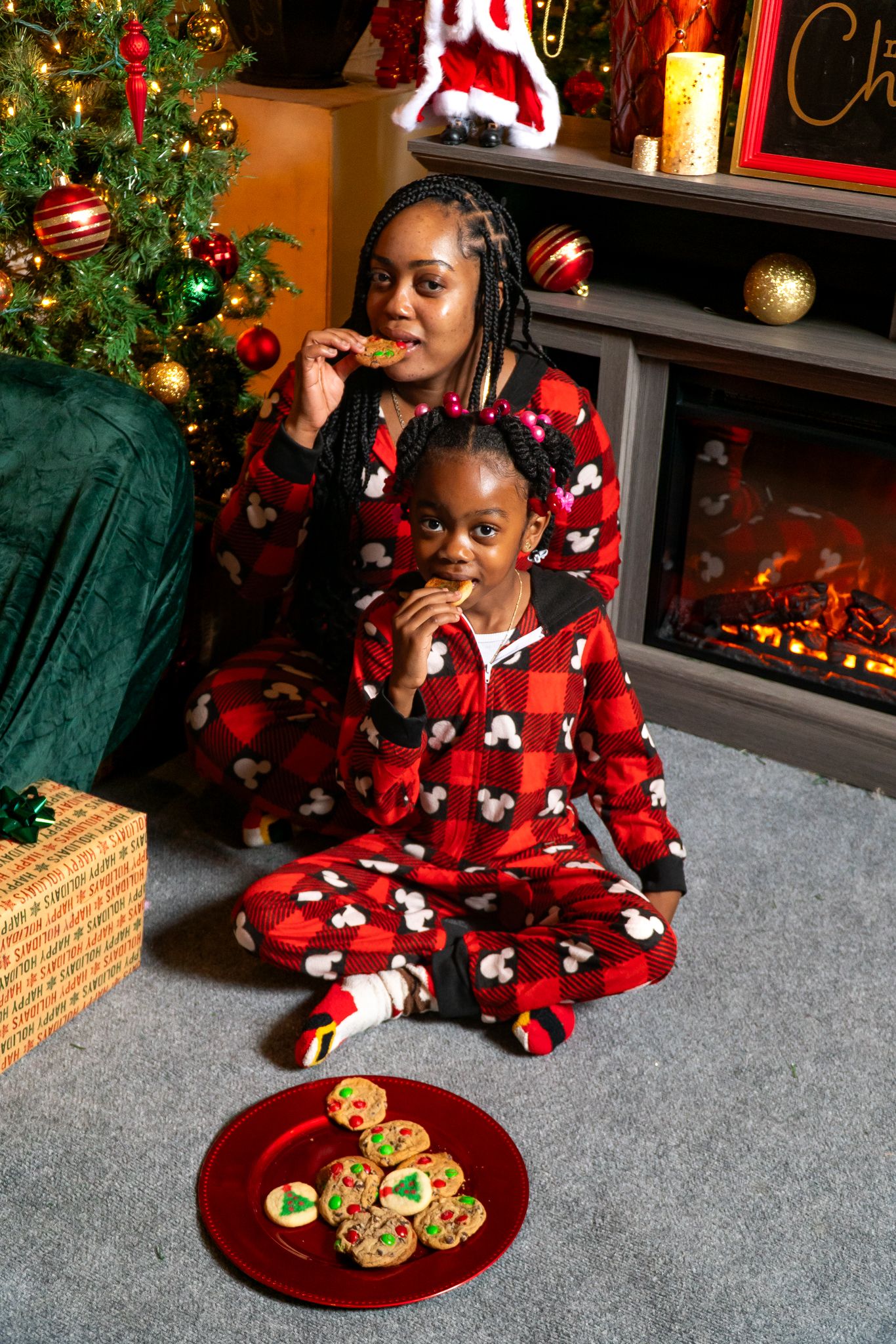 Mother and daughter in matching Mickey Mouse red plaid pajamas eating Christmas cookies by a cozy fireplace.