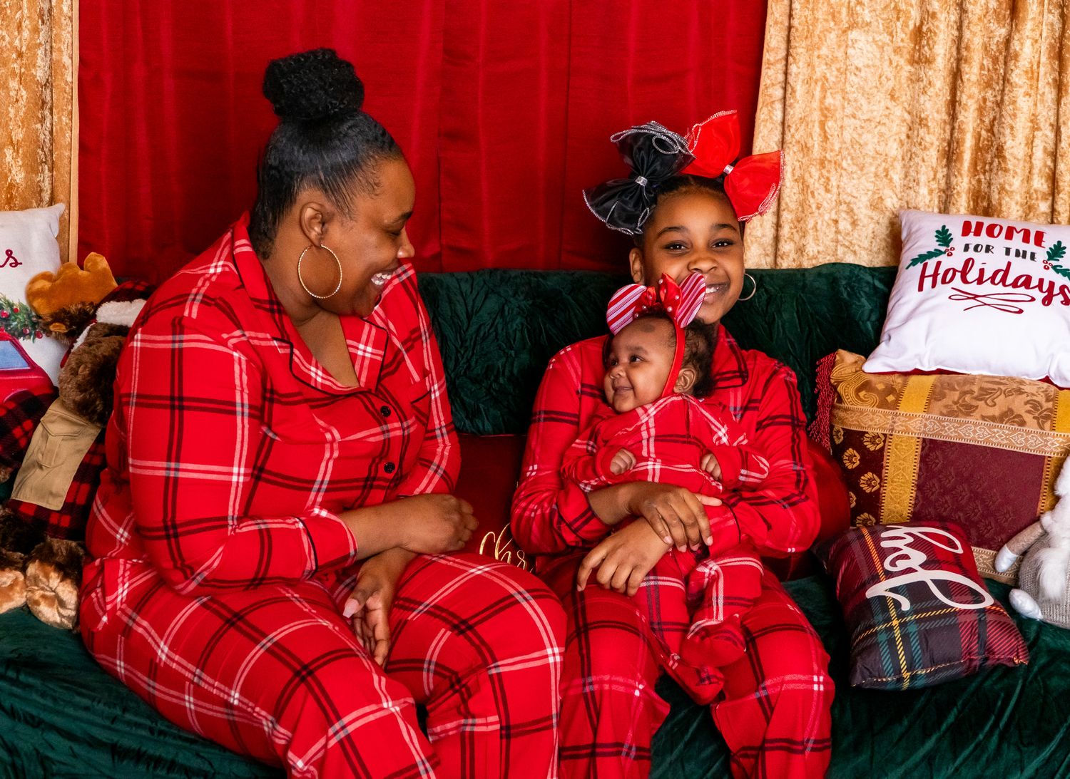 Mother and two daughters in matching red plaid pajamas enjoy a cozy holiday moment on a green velvet sofa.
