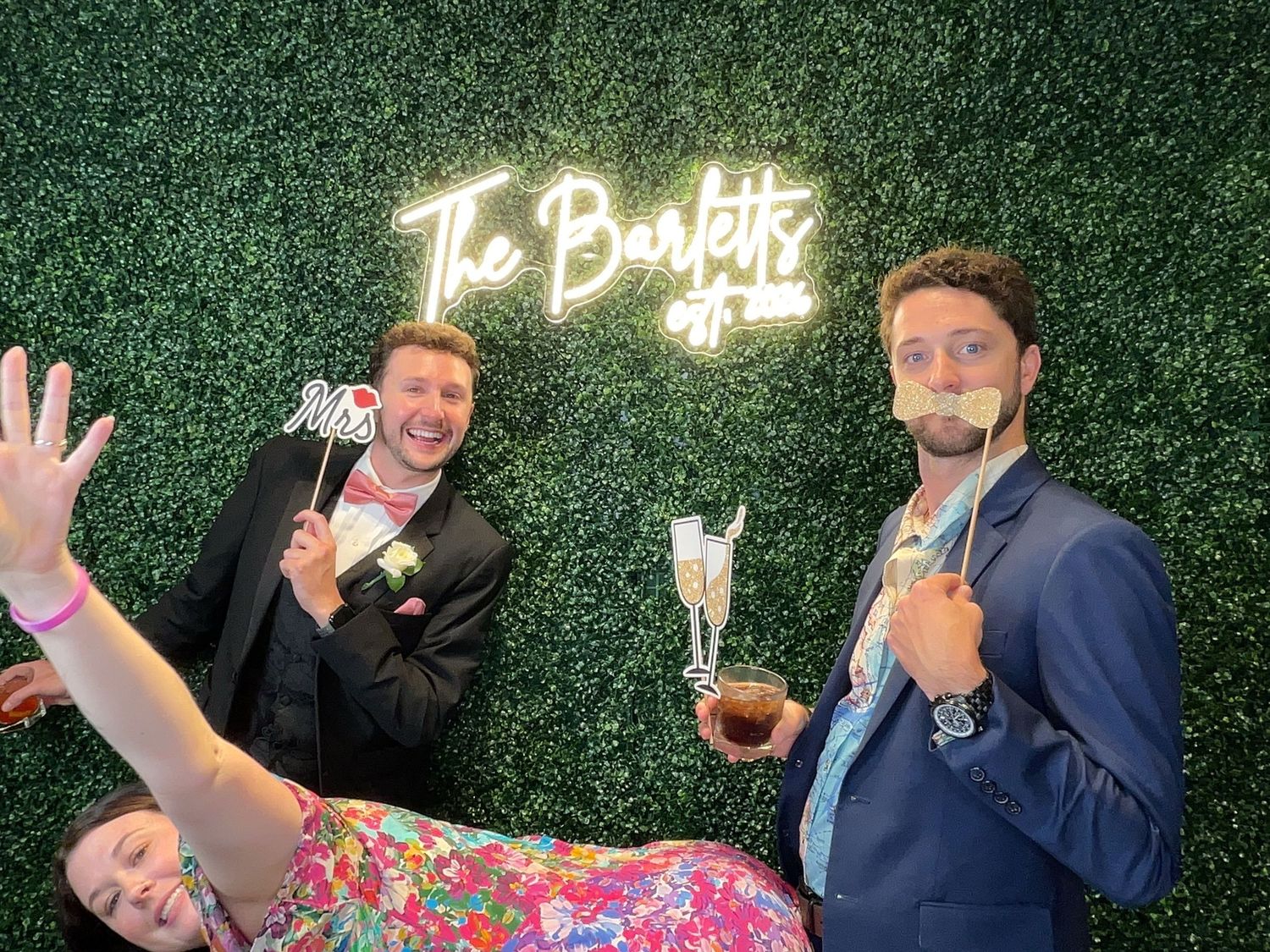 Three guests pose playfully at a wedding photo booth in front of a green hedge wall with a neon sign.